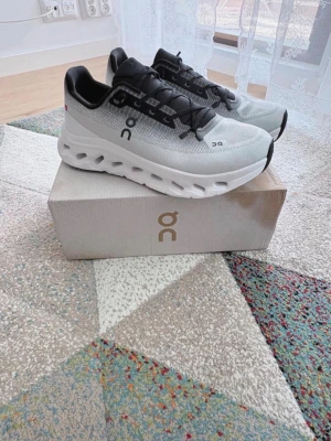 Gråa On Running sneakers med svart detalj - Snygga sneakers från On Running i svarta detaljer och vita, chunky sulor med hålmönster. Skorna har svarta snören och On-logga på sidan. Perfekta för dig som gillar sportig och modern stil.
