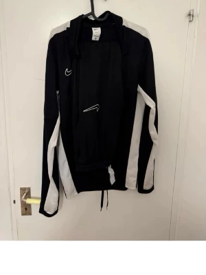 Svart och vit Nike träningsjacka - Snygg svart och vit träningsjacka från Nike med dragkedja framtill och vita detaljer längs ärmarna och axlarna. Jackan har lång ärm och klassisk Nike-logga på bröstet. Perfekt för träning eller sportaktiviteter.