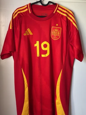 Spaniens matchtröja Lamine Yamal #19 - Röd och gul matchtröja från Adidas för Spaniens landslag med nummer 19 och Lamine Yamal på ryggen. Tröjan har gula detaljer på axlar och sidor, samt landslagets emblem på bröstet. Tillverkad i lätt och ventilerande material, perfekt för fotboll eller träning. som ny väldigt snygg och perfekt till vm! skick 10/10