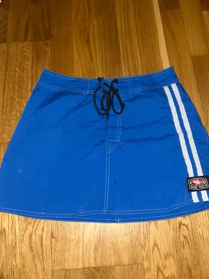 Blå kortkjol från Hot Wave - Sportig blå kortkjol från Hot Wave med vita ränder på sidan och svart snörning i midjan. Kjolen har en patch med logga framtill och är gjord i bomull och polyester. Perfekt för dig som gillar en avslappnad och cool stil.