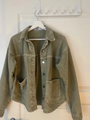 Khaki färg jeansjacka från ZARA - Snygg olivgrön jeansjacka från ZARA med avslappnad passform, stora framfickor och klassisk krage. Jackan har knäppning framtill och långa ärmar med knapp vid ärmslut. Perfekt för en cool och trendig look.