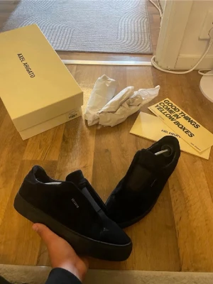 Svarta sneakers från Axel Arigato - Stilrena svarta sneakers från Axel Arigato i mocka med minimalistisk design och diskret logga på sidan. Skorna har en låg siluett och svart sula som ger en clean look. Perfekta för dig som gillar enkel och modern stil.