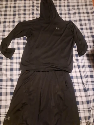 Svart träningsset från Under Armour - Säljer ett svart träningsset från Under Armour med en hoodie och matchande byxor. Hoodien har lång ärm, huva och en stor magficka. Båda plaggen har Under Armour-loggan och är gjorda i ett mjukt, stretchigt funktionsmaterial som passar perfekt till träning.