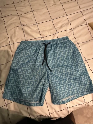 Blå Fendi shorts med logomönster - Säljer ett par blå shorts från Fendi med ikoniskt logomönster över hela plagget. Shortsen har snörning i midjan och är tillverkade i ett lätt material som passar perfekt till sommaren. Snygg och avslappnad passform.