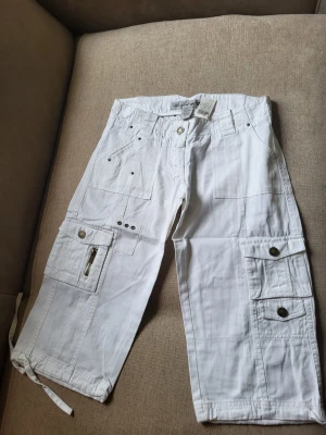 Vita cargobyxor med snörning Wet Seal size 1 USA Xs - Säljer ett par vita cargobyxor med flera fickor, dragkedjedetalj och knappar. Byxorna har snörning vid bensluten och avslappnad passform. Perfekta för en chill och street-inspirerad stil. Materialet är bomull för skön känsla.