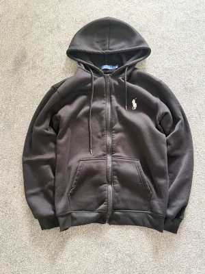Ralph Lauren zipper - Klassisk svart hoodie från Polo Ralph Lauren med dragkedja, två fickor framtill och vit broderad logga på bröstet. Tillverkad i mjukt material och har justerbar huva med snören. Perfekt för en chill och stilren look.