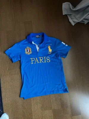 Blå pikétröja från Ralph Lauren  - Snygg blå pikétröja från Ralph Lauren med gul broderad logga och texten PARIS framtill. Tröjan har korta ärmar, krage och knappar vid halsen. Broderade detaljer på bröstet och ärmen ger en exklusiv känsla.