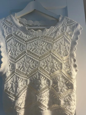 Vit stickad ärmlös topp - Säljer en söt vit stickad ärmlös topp med detaljerat hålmönster i hexagonform. Toppen har rundad halsringning med vågig kant och är gjord i ett mjukt garn, troligtvis bomull. Perfekt för lager på lager är så söt💗💗