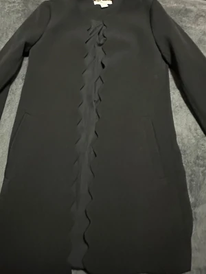 Svart kappa med volangdetalj - Stilren svart kappa med lång ärm och volangdetalj längs hela framsidan. Kappan har två fickor framtill och en rak, klassisk passform. Perfekt för dig som gillar minimalistisk design med en twist.