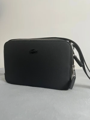 Svart axelväska från Lacoste - Snygg svart axelväska från Lacoste med minimalistisk design och diskret logga framtill. Väskan har justerbar axelrem med Lacoste-tryck och två dragkedjefack. Tillverkad i slitstarkt syntetmaterial med silverfärgade detaljer.