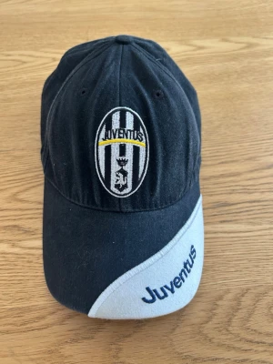 Svart Juventus keps med brodyr - Snygg svart keps från Juventus med klubbens klassiska logga broderad framtill. Skärmen har en vit detalj med blått Juventus-tryck. Perfekt för dig som älskar fotboll och vill visa vilket lag du hejar på. Justerbar baktill för bästa passform.