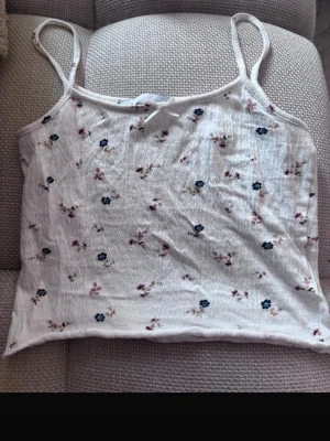Vit blommig linnetopp med rosett - Söt vit linnetopp med små blommor i blått och lila samt en liten rosett framtill. Toppen har tunna axelband och är ärmlös, perfekt för varma dagar. Materialet känns mjukt och lätt, vilket gör den superbekväm att bära.