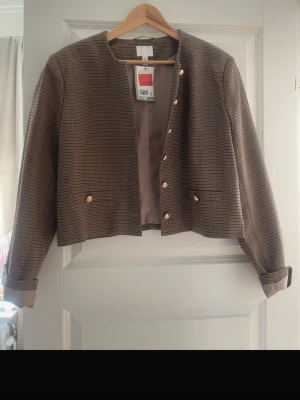 Brunrutig kavaj  - Stilren brunrutig kavaj från H&M med guldfärgade knappar och två fickor framtill. Kavajen har en rak, kortare passform och är helfodrad i beige. Klassisk rundad krage utan slag och uppvikta ärmslut för en modern look.