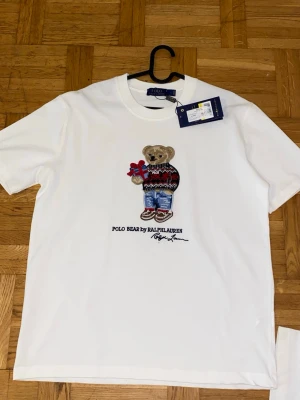 Vit Polo Bear t-shirt från Ralph Lauren - Vit t-shirt från Ralph Lauren med ikonisk Polo Bear-brodyr på bröstet. Björnen har en stickad tröja och jeans, samt håller i ett paket. Klassisk rund hals och korta ärmar. Perfekt för dig som gillar stilrena plagg med en twist.