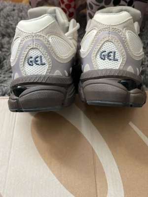 Vita ASICS GEL sneakers - Snygga vita ASICS GEL sneakers med grå och beige detaljer. Skorna har en chunky sula i mörkgrått och meshpaneler för extra ventilation. Klassisk snörning och broderad GEL-logga på hälen. Perfekta för dig som gillar sportig och trendig stil.