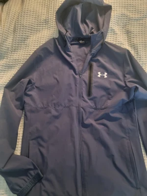 Vindjacka från Under Armour - Snygg vindjacka från Under Armour med huva och hel dragkedja. Jackan har en bröstficka med dragkedja, två sidofickor och justerbara muddar med logga. Perfekt för blåsiga dagar och sportiga outfits. Fast pris. Inga byten. Blågrå färg.
