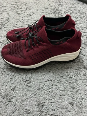 Vinröda sneakers med snabbsnörning - Snygga vinröda sneakers med svart snabbsnörning och vit sula. Ovandelen är i stickad mesh för bra andningsförmåga och komfort. Svarta detaljer på sidan och insidan. Perfekta för dig som vill ha en sportig och modern look.