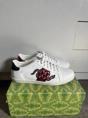 Gucci Ace vita sneakers orm - Säljer ett par vita Gucci Ace sneakers i läder med den ikoniska broderade ormen i rött, svart och grönt på sidan. Skorna har vita snören, gröna och röda detaljer på hälen och klassisk låg profil. Perfekta för dig som vill sticka ut med lyxig streetstyle.