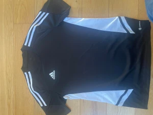 Svart Adidas sporttröja med vita detaljer - Snygg svart sporttröja från Adidas med vita ränder på axlarna och vita paneler på sidorna. Tröjan har korta ärmar och rund hals, samt Adidas-logga på bröstet. I väldigt bra skick