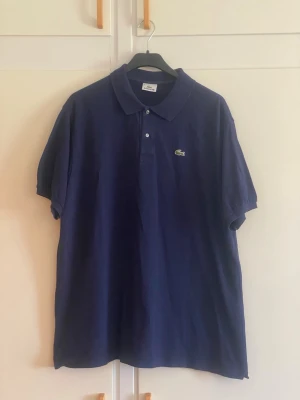 Marinblå pikétröja från Lacoste - Klassisk marinblå pikétröja från Lacoste med korta ärmar, krage och två knappar framtill. Ikoniska krokodilloggan broderad på bröstet. Perfekt för en stilren och sportig look.