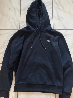 Två hoodies från JJR i svart och grå - Säljer två stilrena hoodies från JJR, en i svart och en i ljusgrå. Båda har huva med dragsko och liten JJR-logga på bröstet. Tillverkade i mjukt sweatshirtmaterial, perfekta för en avslappnad look.