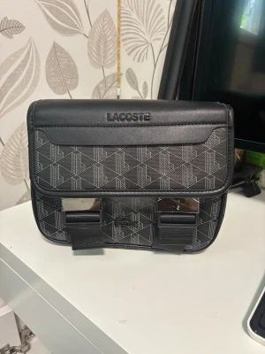 Svart axelväska från Lacoste - Snygg svart axelväska från Lacoste med geometriskt mönster och silverfärgade spännen framtill. Väskan har justerbar axelrem och präglad Lacoste-logga på locket. Tillverkad i konstläder med stilren design som passar till många outfits.