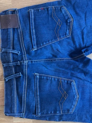 Blå Anbass fit jeans från Replay - Snygga blå jeans från Replay med slim fit passform och klassiska fem fickor. Jeansen har dekorativa sömmar på bakfickorna och ett läderpatch vid linningen. Tillverkade i stretchigt denimtyg för extra komfort. Perfekta till en avslappnad streetstyle-look.