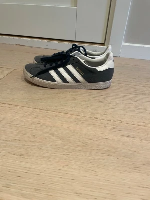 Adidas Gazelle mörkgrå sneakers - Snygga Adidas Gazelle sneakers i mörkgrå mocka med vita detaljer och klassiska tre ränder på sidan. Skorna har svart snörning, vit hälkappa och sula i gummi. Perfekta för dig som gillar retrostil och vill ha något stilrent till vardagslooken.