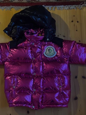 Moncler Genius x Palm Angels “Snow Angels” Tim Jacket in Metallic Pink Nylon - Säljer en helt oanvänd Moncler Genius x Palm Angels Tim Jacket från 8 Moncler Palm Angels-kollektionen, ” Snow Angels ”.  Detta är en exklusiv statement-puffer från Moncler Genius-samarbetet med Palm Angels – en av de mer eftertraktade collab-modellerna. Jackan har blank metallic magenta/lila nylon, svart kontrastdesign och den ikoniska Snow Angels-estetiken, vilket ger den ett väldigt tydligt lyxigt streetwear-uttryck. Det här är alltså inte en vanlig standard-Moncler, utan en runway/collab-pie