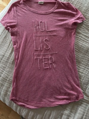 Rosa t-shirt från Hollister - Säljer en rosa t-shirt från Hollister med broderad logga framtill. T-shirten har rund halsringning och korta ärmar. Skön och mjuk bomullskvalitet, perfekt till jeans eller shorts. Små öppningar vid broderier på vissa ställen, se bild