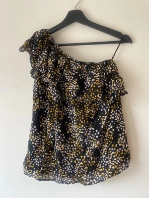 Svart blommig oneshoulder topp - Snygg svart oneshoulder topp med blommigt mönster i vitt och gult. Toppen har volangdetalj vid axeln och är tillverkad i ett lätt, luftigt material som känns perfekt för varmare dagar. Passar dig som vill ha en unik och trendig look. Storlek XS.