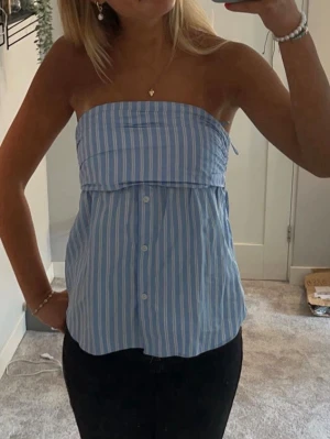 Blåvit randig axelbandslös tubtopp - Snygg blå och vit randig tubtopp med knappar framtill. Toppen är axelbandslös och har en bandeau-formad överdel. Perfekt för dig som vill ha en chill och trendig sommarlook.