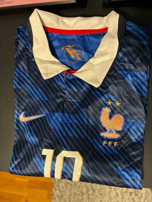 Frankrike Nike fotbollströja blå - Snygg blå fotbollströja från Nike med Frankrikes landslagsmärke och nummer 10 på framsidan. Tröjan har vita och röda detaljer, klassisk krage och två stjärnor ovanför tuppen. Tillverkad i lätt och ventilerande material, perfekt för match eller träning.