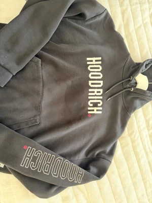 Svart HOODRICH hoodie med tryck - Säljer en svart hoodie från HOODRICH med stor vit logga på bröstet och ärmen. Klassisk känguruficka framtill och justerbar huva med snören. Skön streetwear-stil i mjukt material, perfekt för dig som gillar en avslappnad look.