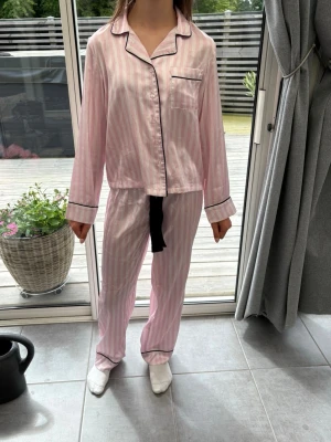 Rosa randig pyjamas med svarta detaljer - Söt pyjamas i rosa med ljusrosa ränder och svarta detaljer. Setet har långärmad skjorta med bröstficka och matchande byxor med rak passform. Materialet är i satin, perfekt för mysiga kvällar hemma.