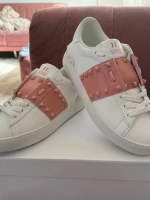 Valentino vita sneakers med rosa nitar - Snygga vita sneakers från Valentino med breda rosa band över vristen dekorerade med matchande pyramidformade nitar. Skorna har klassisk låg profil, vita snören och detaljer i läder. Perfekta för dig som vill sticka ut med en unik och trendig look.