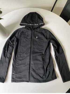 Svart hoodie från Peak Performance - Snygg svart hoodie från Peak Performance med dragkedja framtill och två fickor med dragkedja. Huva med logga på kanten och diskret logotyp på bröstet. Tillverkad i mjukt och stretchigt material, perfekt för en sportig och avslappnad stil.
