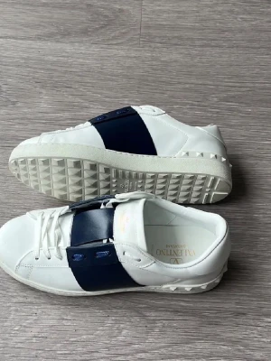 Valentino Garavani vita sneakers - Snygga vita sneakers från Valentino Garavani med bred mörkblå rem över ovandelen. Skorna har en låg silhuett, vita snören och coola nitar på sulan. Tillverkade i slätt läder för en lyxig känsla och unik design.
