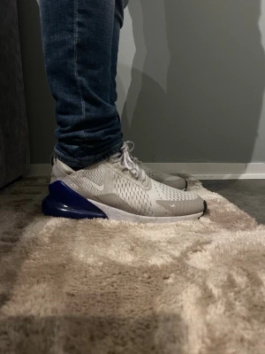 Nike Air Max 270 vita/blå sneakers - Nike Air Max 270 sneakers i vitt och grått med blå häl. Skorna har en luftig mesh-överdel, synlig Air-enhet i hälen och klassisk Nike-logga på sidan. Snygg och sportig design som passar till jeans eller streetwear.