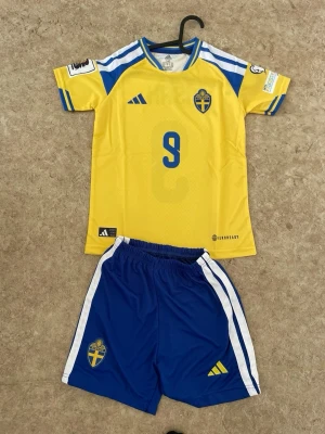 Sverige fotbollströja och shorts Adidas - Gul och blå matchtröja med nummer 9 och namnet ISAK på ryggen, samt blå shorts med vita ränder. Båda plaggen har svenska landslagets emblem och Adidas-logga. Tillverkad i lätt och ventilerande material, perfekt för fotbollsträning eller match.
