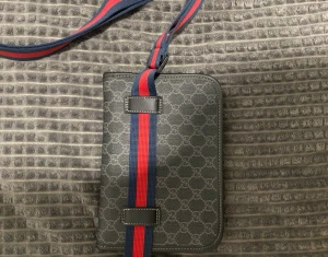  axelväska med monogram och rand - Snygg axelväska från  i svart med klassiskt monogram över hela väskan. Axelremmen är bred i marinblått och rött, vilket ger en sportig touch. Väskan är rektangulär och har detaljer i svart läder. Perfekt för dig som vill ha något trendigt och stilrent.