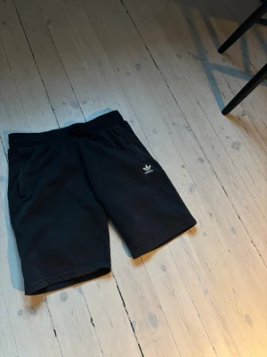 Svarta shorts från Adidas - Svarta shorts från Adidas med klassisk logga på vänster ben. Mjuk resår i midjan och två sidofickor. Perfekta för sommaren och tillverkade i bekvämt bomullsmaterial. Enkel och stilren design som passar till det mesta.