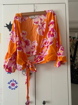 Blommig omlottopp från Monki - Färgstark omlottopp från Monki i orange med stora rosa och vita blommor. Toppen har vida trekvartsärmar och knytband i midjan för en snygg passform. Perfekt för dig som vill sticka ut med en unik och lekfull stil.