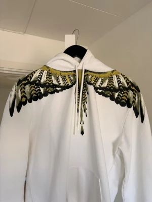 Vit hoodie med fjädermönster från Marcelo Burlon - Säljer en vit hoodie från Marcelo Burlon med unikt fjädermönster i svart, gult och grönt över axlar och bröst. Hoodien har huva med snören och en stor ficka framtill. Materialet är mjukt och bekvämt, perfekt för dig som vill sticka ut med en cool design.