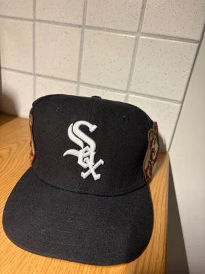 New Era keps - Säljer en svart keps från New Era med White Sox-logga broderad i vitt framtill. På sidan finns en stor brodyr av en ängel i brunt, beige och rött samt text i vitt. Storlek 7 1/2