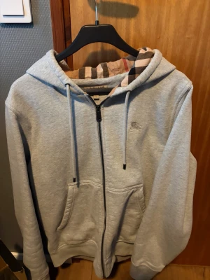 Grå zip up från Burberry - Snygg grå hoodie med dragkedja från Burberry. Klassisk huva med det ikoniska rutmönstret på insidan, dragsnören och två stora fickor framtill. Perfekt för en chill och stilren look. Nyskick använd max 3 gånger. Storlek L men passar mig som är 183cm 75kg.