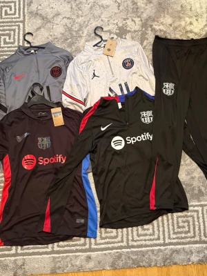 Allt för endast 1500kr - Säljer ett svart träningsset från FC Barcelona med långärmad tröja och byxor. Tröjan har dragkedja vid halsen, röda och blå detaljer samt Spotify-logga och klubbmärke. Tillverkad i ett lätt och snabbtorkande material, perfekt för fotbollsträning.