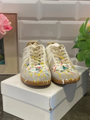 Maison Margiela sneakers med färgstänk - Unika sneakers från Maison Margiela i beige mocka och läder med färgstänk i gult, rött, blått och grönt. Klassisk låg modell med vita snören och gummisula. Perfekt för dig som vill sticka ut med en kreativ och trendig look.