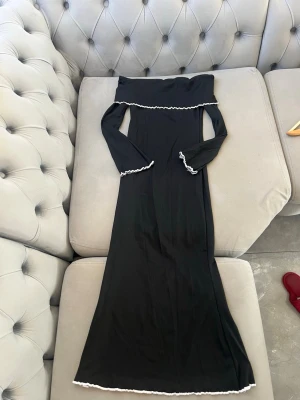 Svart offshoulder långklänning - Trendig svart långklänning med offshoulder-design och långa ärmar. Klänningen har vita volangdetaljer längs kanten upptill, vid ärmsluten och nertill. Tillverkad i ett mjukt, ribbat material som ger en snygg passform och extra komfort.