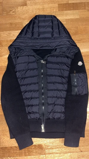 Svart Moncler cardigan dunjacka med huva - Svart hybridjacka från Moncler med dunfylld, quiltad front och huva samt stickade ärmar och rygg. Jackan har dragkedja framtill, en ärmficka med dragkedja och Moncler-logga på ärmen. Perfekt för dig som vill ha en snygg och sportig look. Har dok ett litet hål i fickan men enkelt att fixa.